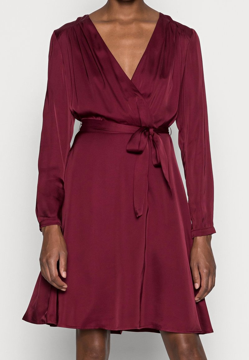 Femme portant une robe portefeuille à manches longues, rouge foncé, avec une ceinture nouée à la taille et un décolleté en V, debout devant un fond uni.