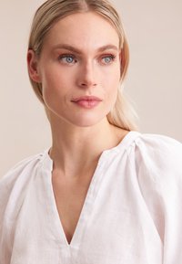 Blusa bianca in lino con scollatura a V, maniche a sbuffo e dettagli di texture sottili. Il tessuto sembra leggero e traspirante.