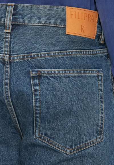 Jeans din denim de culoare albastru mediu, cu cusături contrastante, având un patch pe spate etichetat "FILIPPA K" și un singur buzunar la spate.