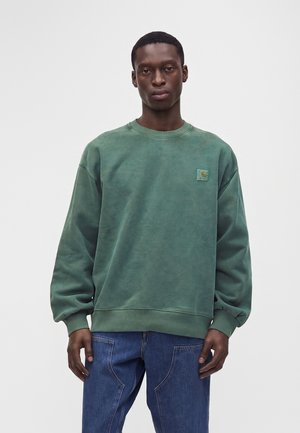 Νέος άνδρας με πράσινο φούτερ Carhartt και μπλε τζιν, στέκεται με ουδέτερη έκφραση μπροστά σε απλό φωτεινό φόντο.