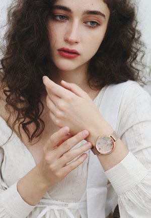 Reloj de oro rosa con una esfera redonda que muestra diseños florales, pulsera de malla de metal y un atuendo blanco texturizado visible en el fondo.