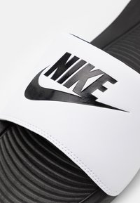 Nike papucs, fehér műanyag pánttal, fekete logóval és texturált fekete gumitalppal. Állítható méret, könnyű kialakítás.