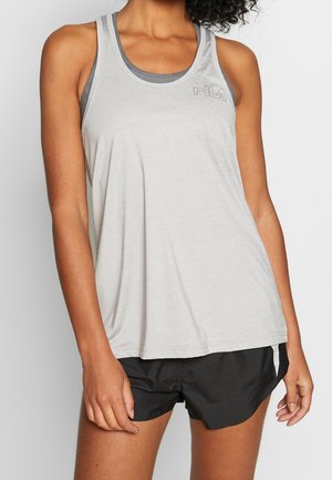 Femme portant un débardeur d'entraînement FILA gris clair par-dessus un soutien-gorge de sport gris foncé et un short de sport noir, debout devant un fond blanc.