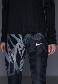 Maglietta nera a maniche lunghe abbinata a leggings neri con un motivo geometrico astratto grigio e un logo Nike bianco sulla vita.
