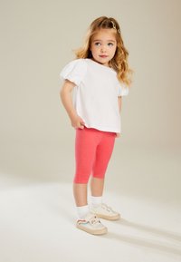 Witte katoenen t-shirt met pofmouwen, gecombineerd met roze leggings en crèmekleurige schoenen met een regenboogontwerp. De achtergrond is neutraal.