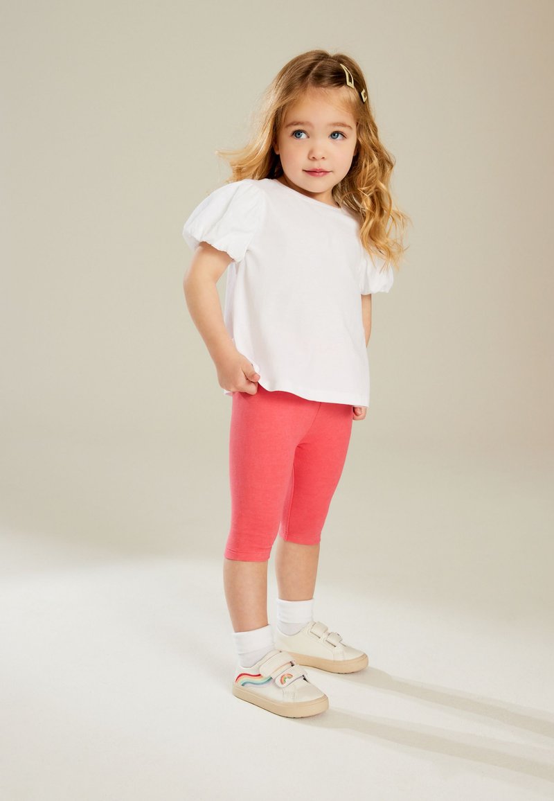 Witte katoenen t-shirt met pofmouwen, gecombineerd met roze leggings en crèmekleurige schoenen met een regenboogontwerp. De achtergrond is neutraal.