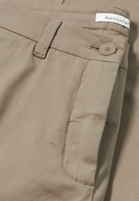 Pantalons en coton beige avec une texture lisse. Les caractéristiques incluent une fermeture à bouton, des détails de poches cousus et une étiquette sur la taille.
