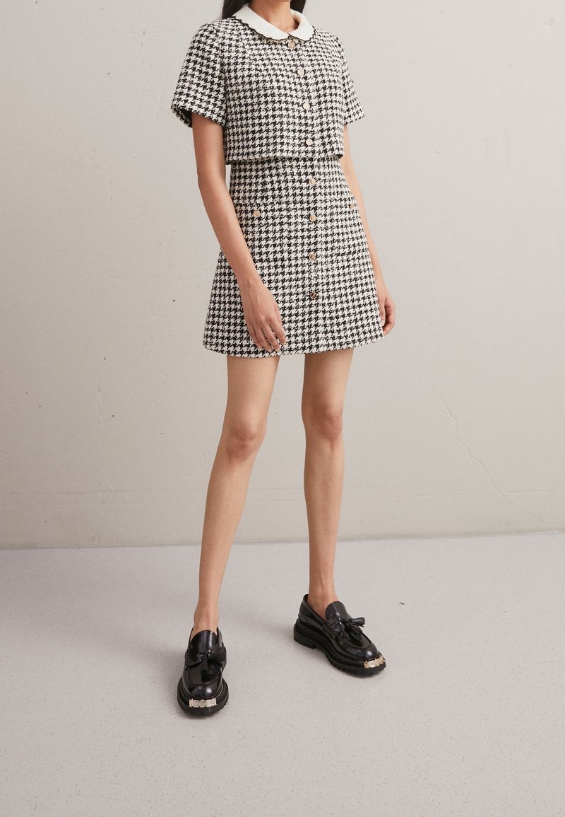 Femme portant une robe à manches courtes motif pied-de-poule noir et blanc, avec col blanc et boutons, associée à des mocassins noirs, debout à l'intérieur.