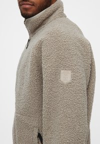Beige fleece pullover med høj krave, med tekstureret stof, sidelommer med lynlås og et logo-patch på ærmet.