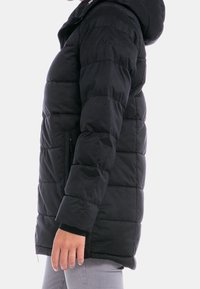 Svart puffjacka med huva, som har en quiltad design, framfickor och logodetalj på axeln. Mjuk, något glänsande textur.