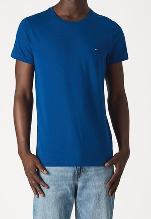 Blaues Baumwoll-T-Shirt mit rundem Ausschnitt und kurzen Ärmeln. Mit einem kleinen Logo auf der linken Brust. Kombiniert mit hellblauen Jeans.