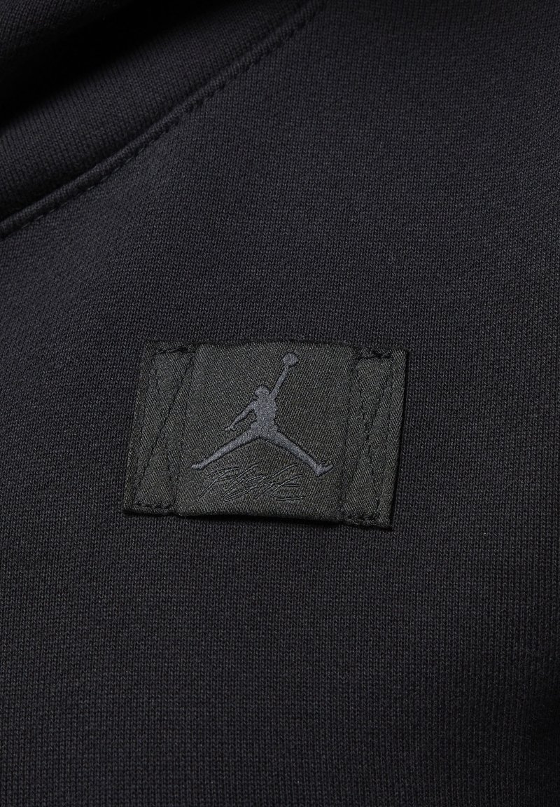 Il materiale della felpa nera presenta un patch testurizzato con il logo Jumpman grigio scuro e la firma, cucito con bordi precisi.