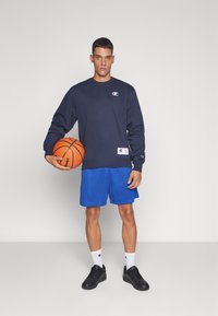 Sudadera navy con logo, pantalones cortos de malla azules y zapatillas negras. El modelo sostiene un balón de baloncesto naranja, de pie contra un fondo liso.