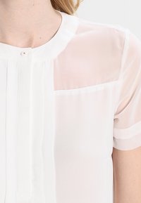 Blusa bianca a maniche corte con colletto, che presenta una parte superiore trasparente e una chiusura con bottoni. Plissettature verticali aggiungono texture al tessuto.