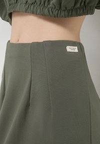 Gonna verde oliva realizzata in tessuto morbido, con pieghe, vita aderente e un piccolo etichetta che recita "DESIGN ZERO WASTE" sul retro.