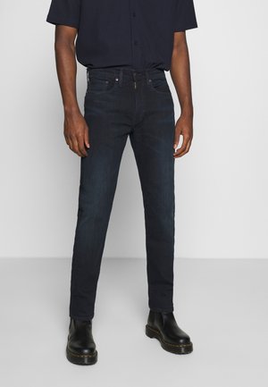 Mann trägt dunkelblaue Slim-Fit-Jeans, ein schwarzes Kurzarmshirt und schwarze Lederstiefel und steht vor einem schlichten, hellen Hintergrund.