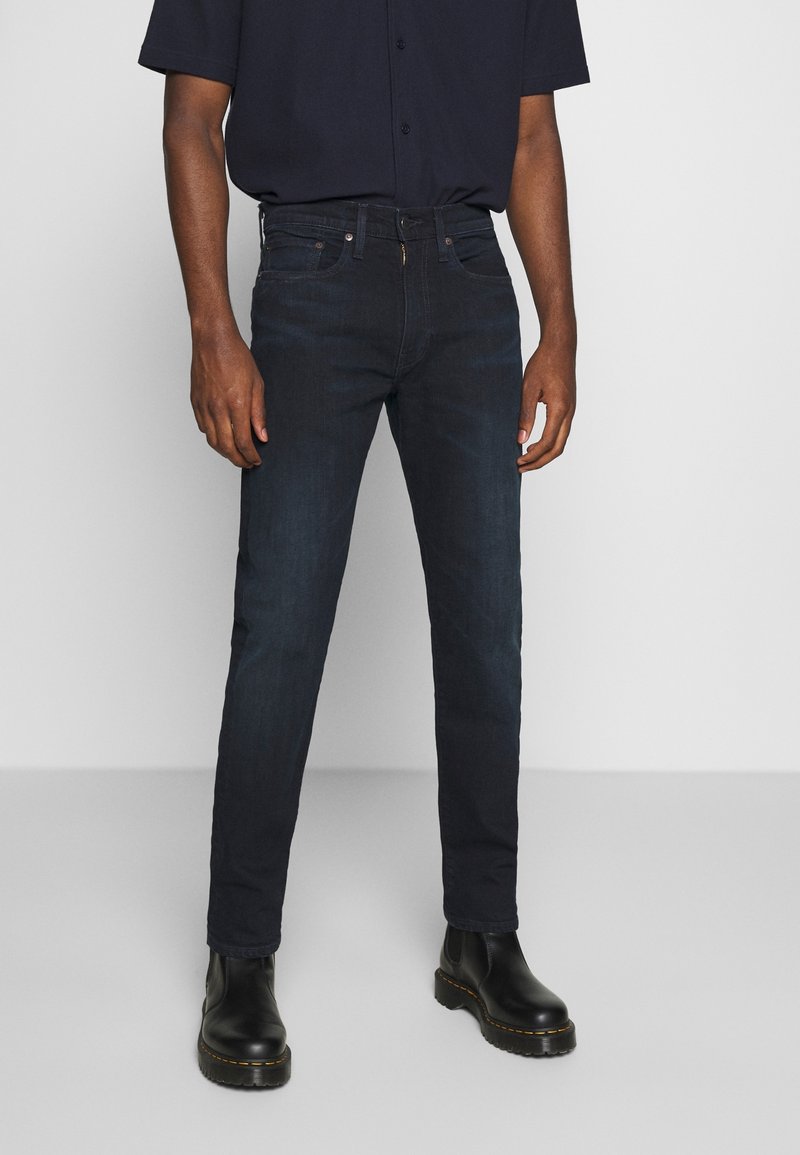 Homme portant un jean slim bleu foncé, une chemise noire à manches courtes et des bottes en cuir noires, debout devant un fond uni et clair.