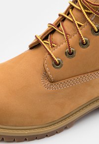 Scarponcino da trekking in suede marrone con lacci gialli e borgogna, occhielli metallici, cuciture bianche e suola in gomma testurizzata.