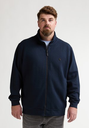 Marineblaue Reißverschlussjacke aus strukturiertem Stoff mit geometrischem Muster, ausgestattet mit einem hohen Kragen und gerippten Bündchen. Einfaches Logo-Detail auf der Brust.