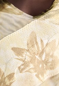 Svetložltý textil s kvetinovými vzormi a textúrovaným tkaným povrchom, doplnený priesvitnou sieťovinou pozdĺž výstrihu.