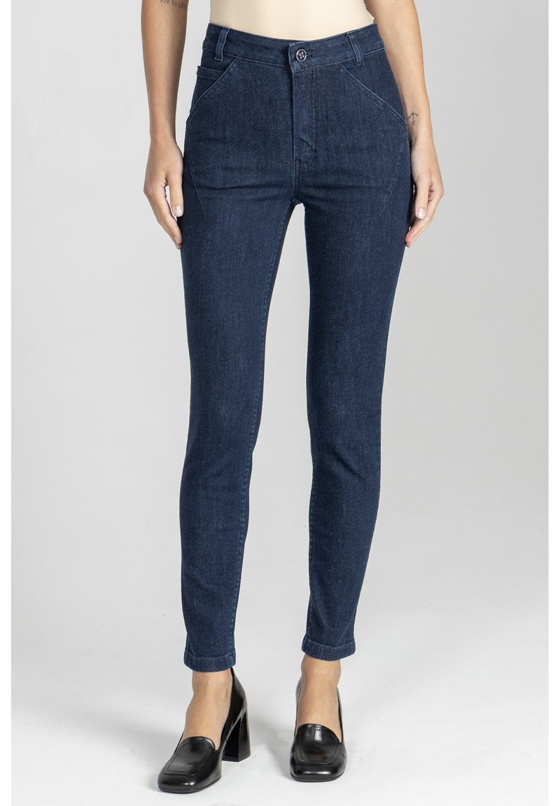 Kobieta w ciemnoniebieskich dopasowanych dżinsach i czarnych mokasynach na obcasach, stojąca na jednolitym jasnoszarym tle.