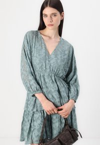 Lyseblå blomstret blondekjole med lange ermer, V-hals, rynket midje og lagdelt skjørt. Holdt med en mørk brun veske.