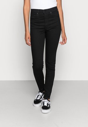 Persoon draagt zwarte skinny jeans met hoge taille en zwart-witte veter sneakers, staand tegen een effen lichtgrijze achtergrond.