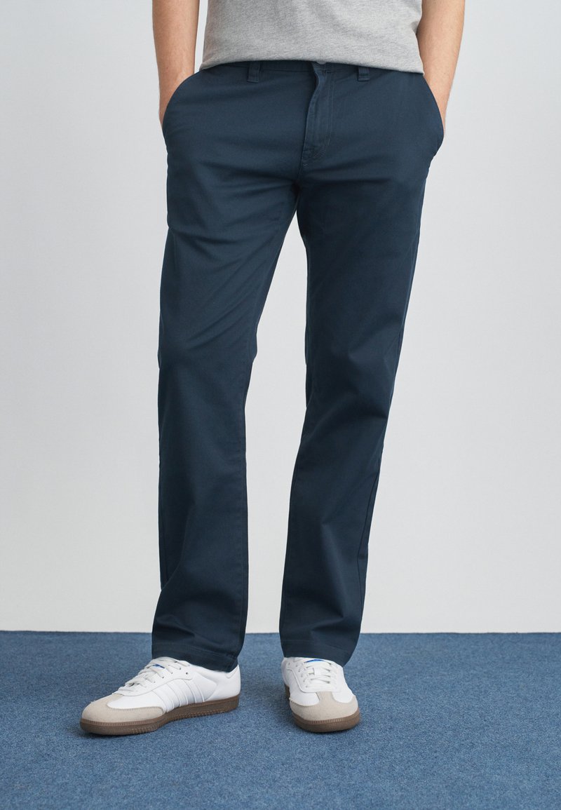 Ναυτικό μπλε βαμβακερά chino με ίσια γραμμή, με πλαϊνές τσέπες. Συνδυασμένα με λευκά αθλητικά παπούτσια και γκρι μπλουζάκι. Απλός σχεδιασμός.