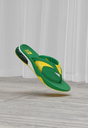 Sandale sport verde și galben tip flip-flop cu talpă căptușită și branț texturat, plutind deasupra podelei din lemn deschis și a unui fundal cu perdea albă.