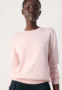 Lys rosa genser laget av mykt stoff. Har rund hals, lange ermer, og en liten brodert logo på brystet.
