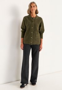 Grüner, flauschiger Cardigan mit fünf Knöpfen, Raglanärmel und gerippten Bündchen, kombiniert mit dunklen, weit geschnittenen Jeans und schwarzen Turnschuhen.