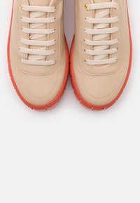 Baskets en cuir beige avec semelles en caoutchouc orange, dotées de lacets plats beiges et d'œillets dorés. Texture lisse avec des détails de coutures courbées.