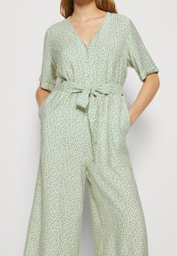 Mono verde con estampado, de mangas cortas, escote en forma de v, cinturón en la cintura y bolsillos laterales. La tela tiene una textura suave y liviana.