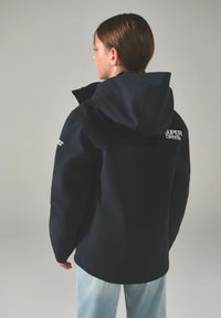 Marinblå jacka med huva, med svart axelpanel och en vit logotyp. Slät textur med en avslappnad passform, matchad med ljusblå jeans.