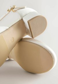 Witte laag-gehakte sandalen met enkelband en gouden gesp, met zichtbare gestructureerde bruine rubberen zolen en blokhak.
