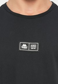Czarny t-shirt z krótkim rękawem z okrągłym dekoltem oraz prostokątną metką z logo Kappa z białym napisem po lewej stronie.