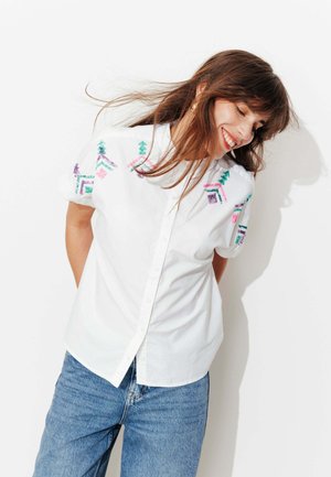 Femme souriante aux cheveux bruns portant une chemise blanche à manches courtes avec une broderie géométrique colorée et un jean bleu, debout devant un fond blanc.