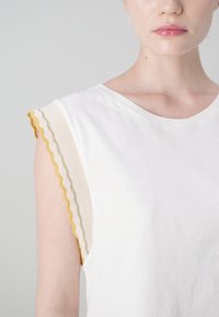 Hvit t-skjorte med rund hals, med bølgete detaljer i beige og gult på ermene, laget av mykt bomullsmateriale.