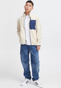 Fleecejakke i kremfarge med en marineblå brystlomme, hel glidelås og høy krage; parret med blå jeans og hvite joggesko med svarte striper.