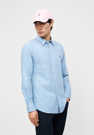 Young man wearing a light blue checkered button-up shirt, dark pants, and a light pink baseball cap, standing with one hand in pocket.