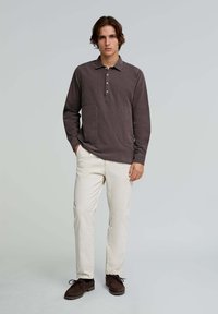 Long-sleeve, brown cotton shirt with a collared neckline, button placket, and front pocket, paired with cream-colored pants and brown shoes.