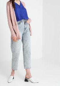 Rosa stickad cardigan, blå blus, ljus tvätt med hög midja jeans och vita skor med svarta klackdetaljer. Släta texturer och lagerdesign.