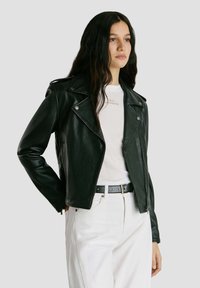 Kvinde iført sort læderjakke over hvid t-shirt og hvide bukser med sort bælte, stående mod en lysegrå baggrund.