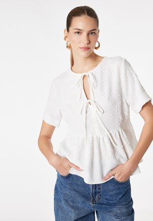 Młoda kobieta z gładko zaczesanymi włosami związanymi z tyłu nosi złote kolczyki koła, białą fakturowaną bluzkę z krótkim rękawem i wiązaniami z przodu oraz niebieskie jeansy, ręce w kieszeniach.