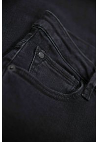 Jean en denim noir avec une poche avant dotée d'un bouton visible. Le tissu a une texture lisse et des accents de coutures subtils.