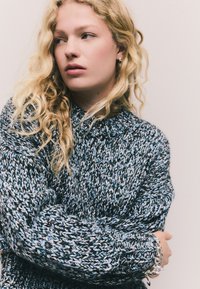 Suéter de punto grueso en tonos negros y azules con cuello alto, patrón texturizado y puños acanalados, combinado con una pulsera plateada.