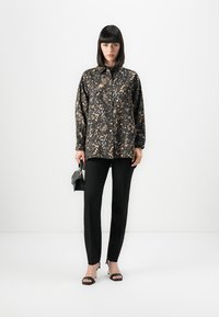 Donna che indossa una camicia nera e beige a fantasia, leggera e con bottoni, pantaloni neri aderenti, tacchi aperti neri, mentre tiene una piccola borsa nera.