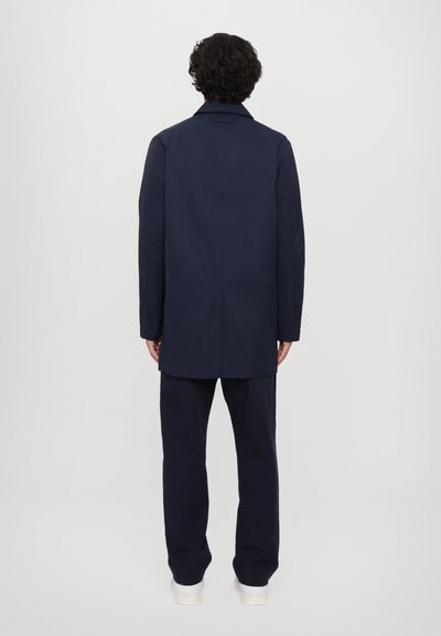 Manteau bleu marine à coupe droite, à manches longues et avec un col en V. Porté sur un pantalon bleu marine assorti et des chaussures de couleur claire.