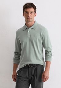Polo cu mânecă lungă de culoare verde mentă, cu guler, având un sistem de închidere cu trei butoane și o detaliere subtilă a logo-ului pe piept. Asortat cu pantaloni gri.