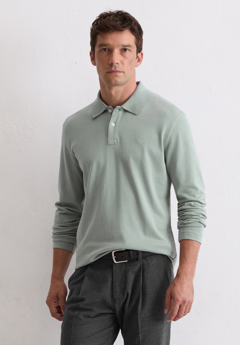Polo cu mânecă lungă de culoare verde mentă, cu guler, având un sistem de închidere cu trei butoane și o detaliere subtilă a logo-ului pe piept. Asortat cu pantaloni gri.